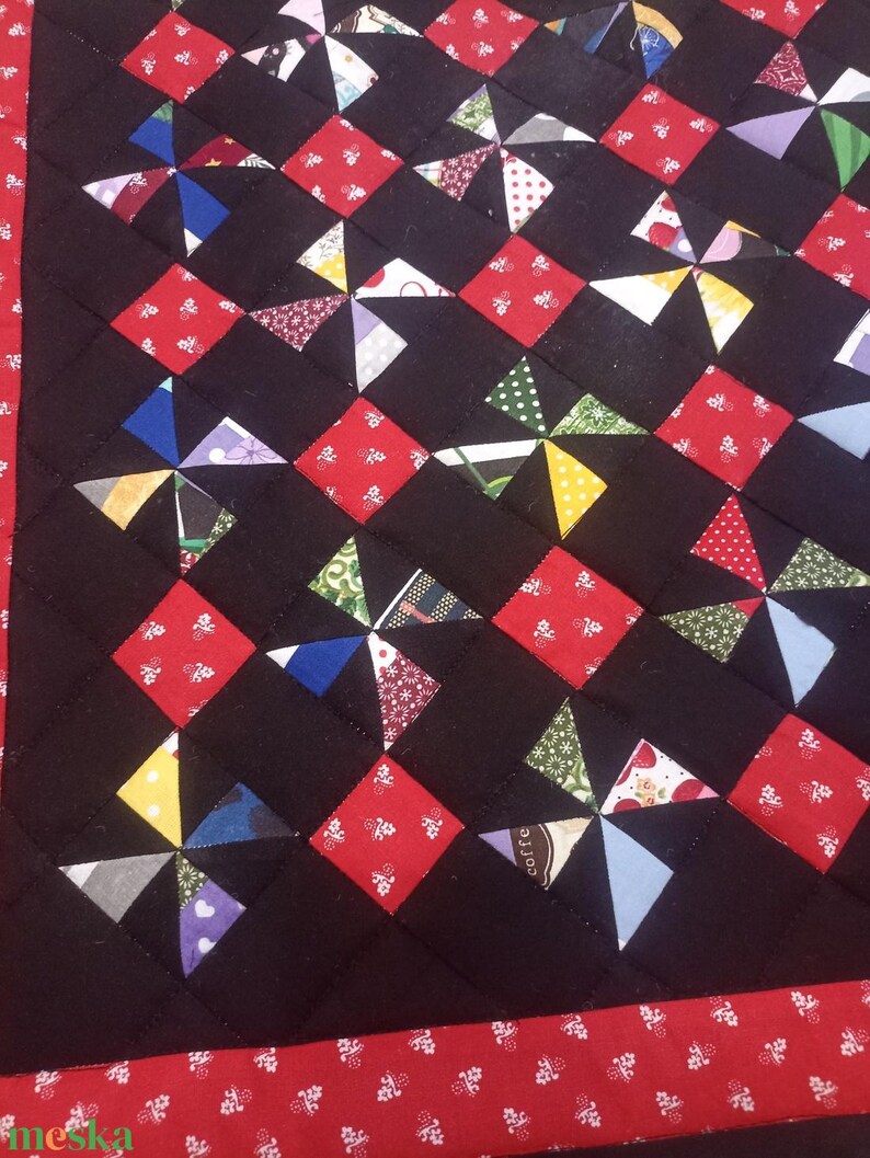 May include: Close-up of a handmade quilt with a black background. The quilt features a repeating pattern of red squares and colourful pinwheel designs. The border is red with a white floral pattern.