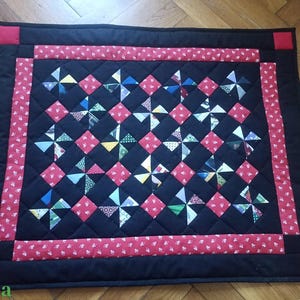 May include: A handmade quilt with a black background and a red border. The quilt features a pattern of red squares and pinwheel designs in various colours. The corners have red squares. The quilt is likely made of cotton.