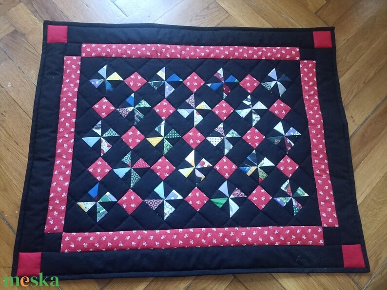 May include: A handmade quilt with a black background and a red border. The quilt features a pattern of red squares and pinwheel designs in various colours. The corners have red squares. The quilt is likely made of cotton.