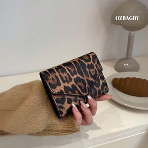 May include: A small, rectangular wallet with a leopard print pattern. The wallet has a flap closure and is black on the sides and back. The text "OZBAGBY" is visible in the background.