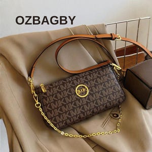 Puede incluir: Bolso de hombro marrón con un patrón repetido de las letras "M" y "K". El bolso tiene una correa de cadena dorada y una correa de cuero marrón. El texto "OZBAGBY" es visible en la parte superior.