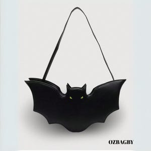 May include: A black bat-shaped handbag with a long, thin strap. The bag has a detailed bat silhouette with pointed ears and wings. The bat's eyes are green. The text "OZBAGBY" is at the bottom.