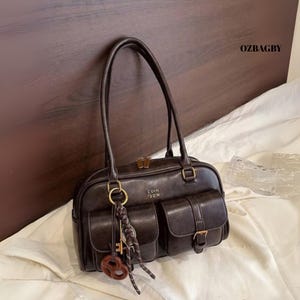 Puede incluir: Un bolso de mano de cuero marrón oscuro con dos bolsillos delanteros, herrajes dorados y un charm en forma de llave. El bolso tiene dos asas y el texto "COIH 120N" es visible en la parte delantera. La marca "OZBAGBY" está impresa en la parte superior derecha.