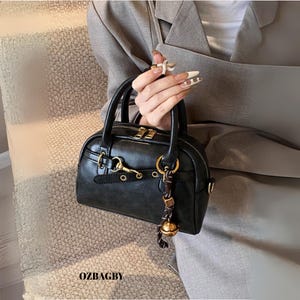 May include: A small, black handbag with a rounded shape and top handles. The bag features gold-coloured hardware, including a zip, clasps, and a decorative charm with a bell. The text "OZBAGBY" is visible at the bottom.