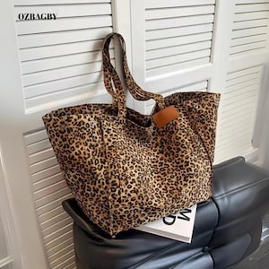 May include: A large tote bag featuring a leopard print design. The bag has two handles and a brown leather detail. The bag is placed on a black leather chair. The text "OZBAGBY" is visible in the top left corner.