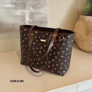 May include: A brown tote bag with a floral and bow pattern. The bag has brown handles and trim, with a gold accent on the front. The text "OZBAGBY" is printed on the bottom left of the bag.