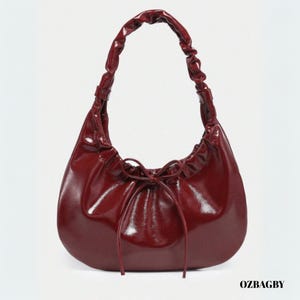 Può includere: Una borsa a mano bordeaux lucida con manico arricciato e chiusura a coulisse. La borsa ha una forma arrotondata e una finitura lucida. Il testo "OZBAGBY" è visibile in basso a destra.