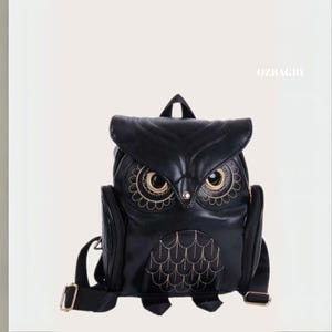 Może przedstawiać: Czarny plecak w kształcie sowy ze złotymi akcentami. Plecak ma szczegółowy design z wyszywanymi wzorami i górnym uchwytem. Oczy są w kolorze złotym, a tekst "OZBAGBY" jest widoczny po prawej stronie.