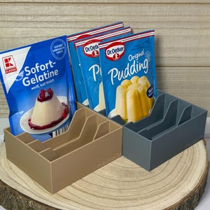 Könnte beinhalten: Zwei rechteckige Kunststoff-Organizer, einer in Beige und einer in Grau, jeweils mit internen Trennwänden. Die Organizer enthalten mehrere Lebensmittelbeutel, darunter eine blaue Kaufland Sofort-Gelatine-Packung und Dr. Oetker Pudding-Tüten.