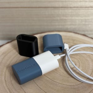 Pode incluir: Um adaptador de cabo de carregamento branco e azul com o texto "PrintFind3D" impresso, juntamente com duas tampas separadas, uma preta e outra azul. Um cabo de carregamento branco também está presente. Os itens estão em uma superfície de madeira.