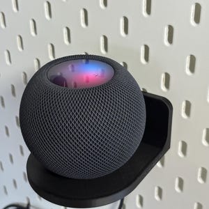 Op de afbeelding: Een donkergrijze, bolvormige slimme luidspreker met een buitenkant van mesh-stof. De bovenkant van de luidspreker heeft een kleurrijk, verlicht display. Hij staat op een zwarte, gebogen plank die aan een witte pegboard-muur is bevestigd.