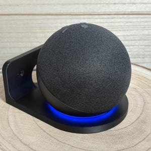 Op de afbeelding: Een zwarte slimme luidspreker met een stoffen hoes staat op een zwarte standaard met blauw licht. De luidspreker is rond en heeft knoppen aan de bovenkant. De standaard staat op een houten oppervlak.