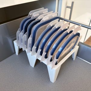 Può includere: Un organizer in plastica bianca contiene diversi coperchi blu e trasparenti per la conservazione degli alimenti. I coperchi sono ordinatamente disposti in posizione verticale, con un divisore in plastica bianca. L'organizer è su un piano di lavoro grigio.