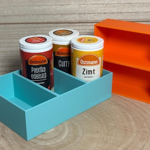 Könnte beinhalten: Ein hellblauer Gewürzorganizer aus Kunststoff mit drei Fächern, der drei zylindrische Gewürzdosen mit den Aufschriften Ostmann Paprika Edelsüß, Curry und Zimt enthält. Daneben steht ein leeres, orangefarbenes Kunststoffregal mit zwei Ebenen.