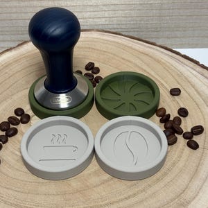 Könnte beinhalten: Ein Kaffeetampfer mit dunkelblauem Griff und Edelstahlbasis sowie zwei grüne und zwei hellgraue Untersetzer zum Thema Kaffee. Kaffeebohnen sind um die Artikel herum verstreut, alles auf einer Holzoberfläche präsentiert.