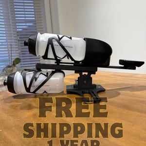 Puede incluir: Dos botellas de agua transparentes con tapas negras y jaulas negras, montadas en un soporte negro. El soporte está sobre una superficie de madera. El texto "FREE SHIPPING 1 YEAR WARRANTY" es visible.