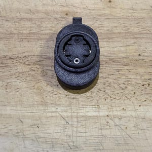 May include: A black 3D-printed bicycle computer mount. The mount has a circular design with a central opening and a small tab at the top. It is designed to hold a cycling computer and is made of a textured material.