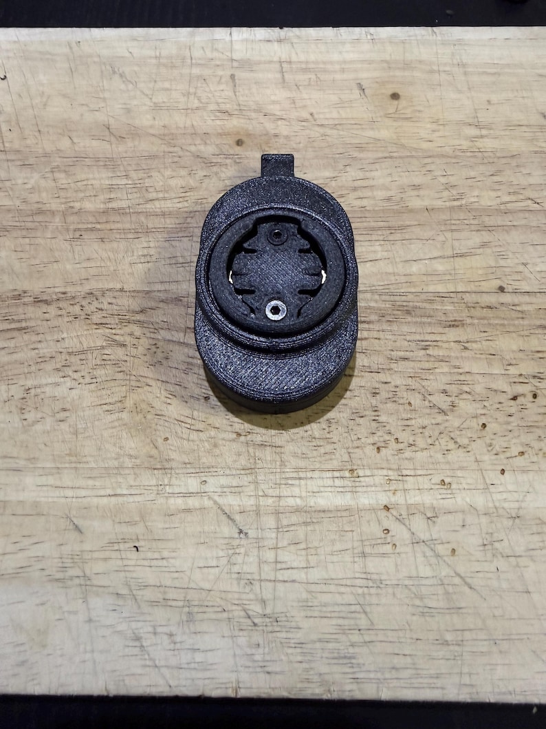 May include: A black 3D-printed bicycle computer mount. The mount has a circular design with a central opening and a small tab at the top. It is designed to hold a cycling computer and is made of a textured material.