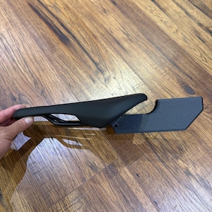 May include: A black bicycle saddle with a streamlined design. The saddle is positioned next to a black metal bracket. The saddle is designed for comfort and performance. The saddle is made of a durable material.