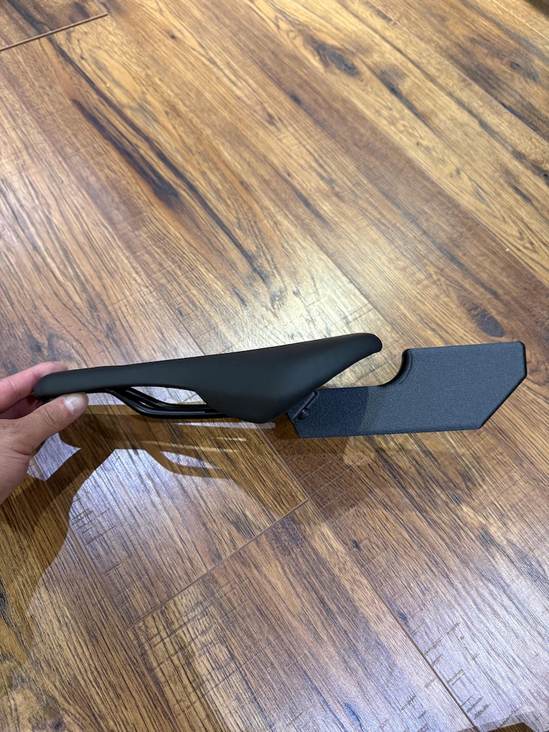 May include: A black bicycle saddle with a streamlined design. The saddle is positioned next to a black metal bracket. The saddle is designed for comfort and performance. The saddle is made of a durable material.