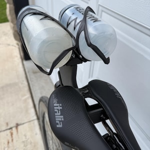 May include: A black bicycle saddle with the Selle Italia logo and two water bottles in black cages. The bottles are clear and have black text. The bike is mounted on a black frame.