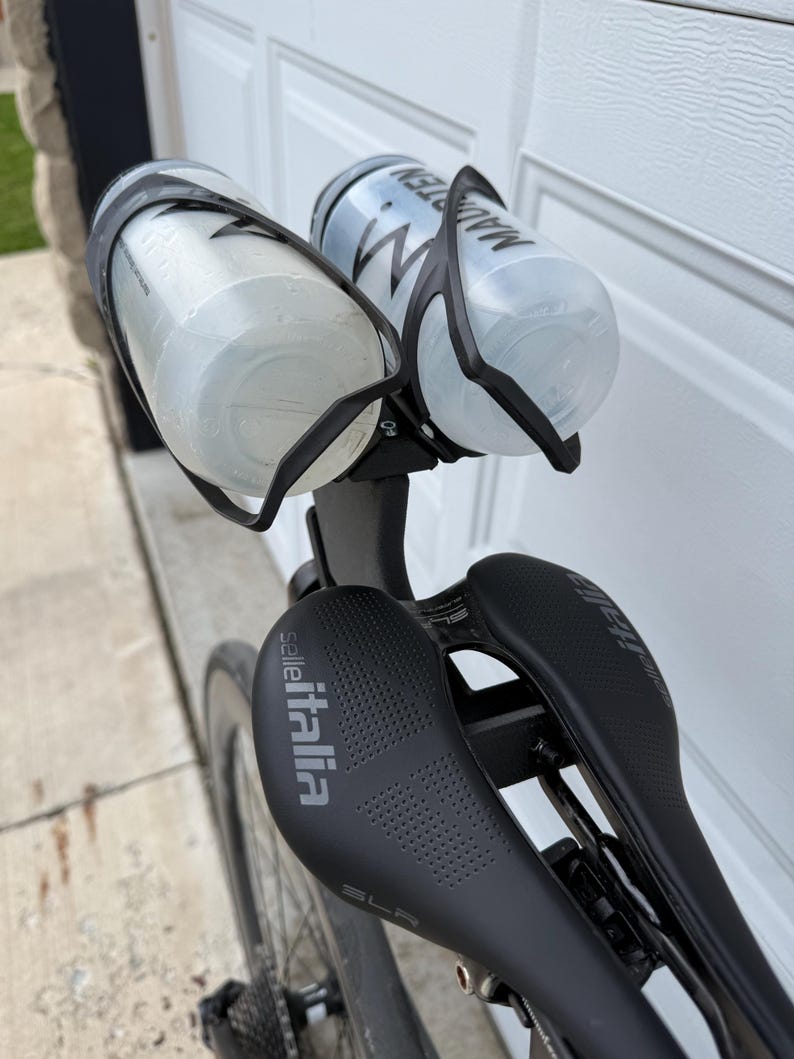 May include: A black bicycle saddle with the Selle Italia logo and two water bottles in black cages. The bottles are clear and have black text. The bike is mounted on a black frame.
