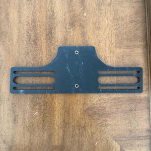 May include: A black metal bracket with a rectangular shape and three horizontal slots on each side. Two small holes are visible near the top. The bracket is placed on a wooden surface.