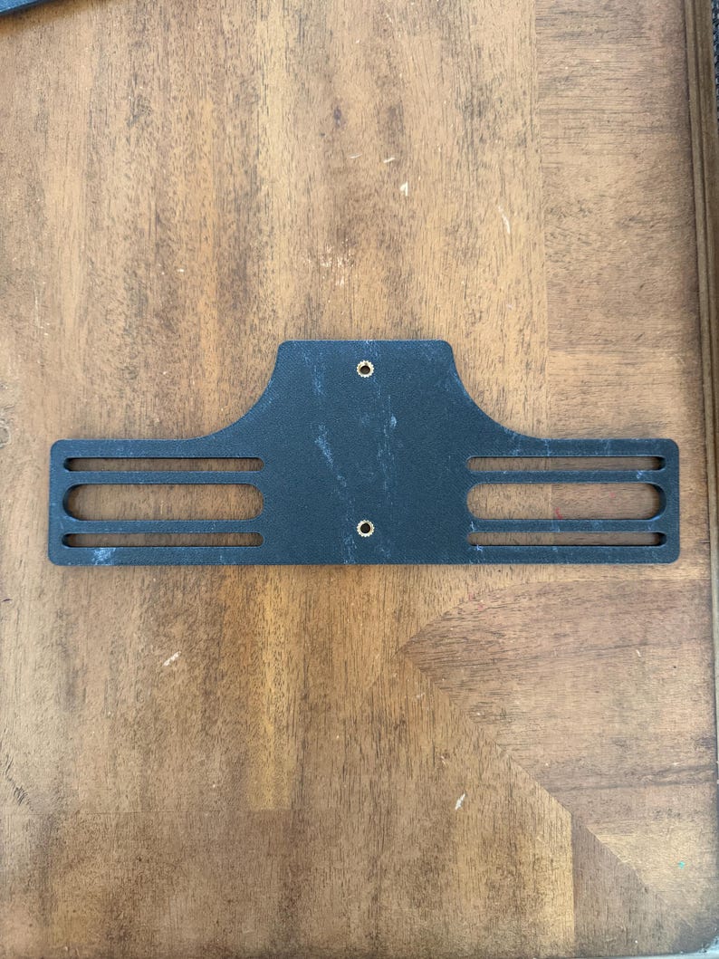 May include: A black metal bracket with a rectangular shape and three horizontal slots on each side. Two small holes are visible near the top. The bracket is placed on a wooden surface.
