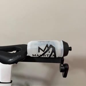 Puede incluir: Una botella de agua transparente con tapa negra y la palabra "MAURTEN" en letras negras. La botella est&aacute; montada en un soporte negro sujeto a una bicicleta. El sill&iacute;n de la bicicleta es visible.