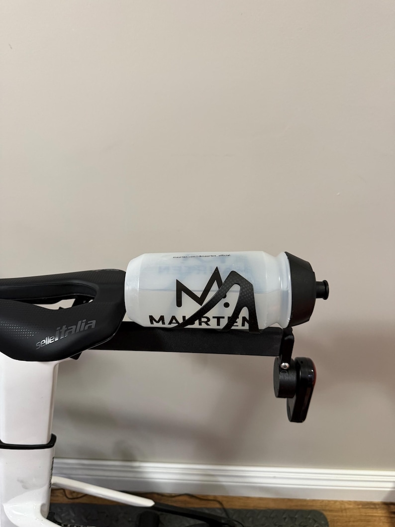 Puede incluir: Una botella de agua transparente con tapa negra y la palabra "MAURTEN" en letras negras. La botella est&aacute; montada en un soporte negro sujeto a una bicicleta. El sill&iacute;n de la bicicleta es visible.