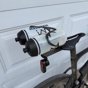 May include: A black bicycle with two water bottles attached to the seat. The bottles are white and black with the word "MAGRTEN" printed on them. A red rear light is mounted below the bottles.
