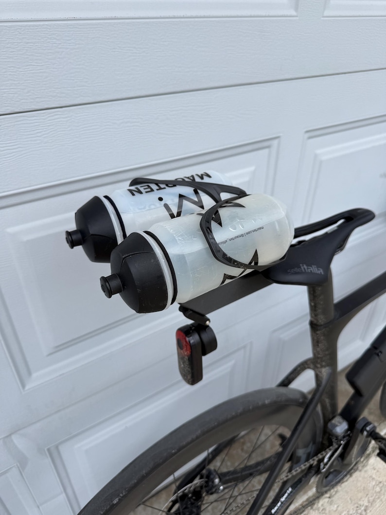 May include: A black bicycle with two water bottles attached to the seat. The bottles are white and black with the word "MAGRTEN" printed on them. A red rear light is mounted below the bottles.