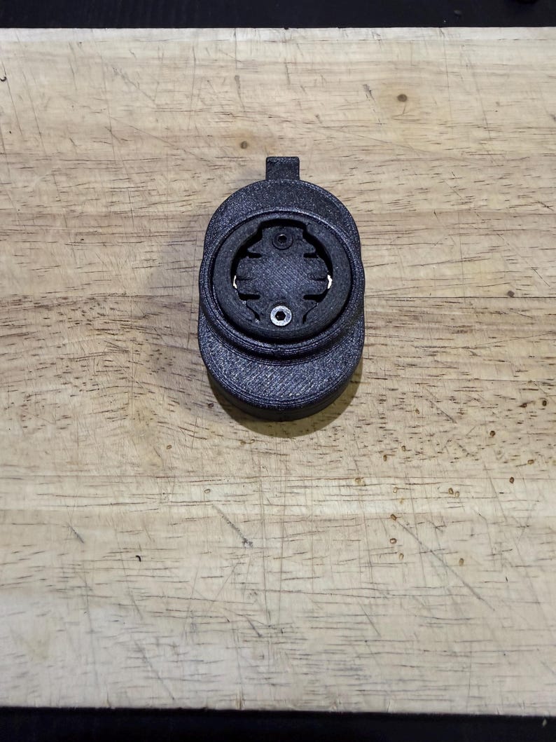 Puede incluir: Un soporte negro para ordenador de bicicleta, impreso en 3D. El soporte tiene un dise&ntilde;o circular con una abertura central y una peque&ntilde;a pesta&ntilde;a en la parte superior. Est&aacute; dise&ntilde;ado para sujetar un ordenador de ciclismo y est&aacute; hecho de un material texturizado.