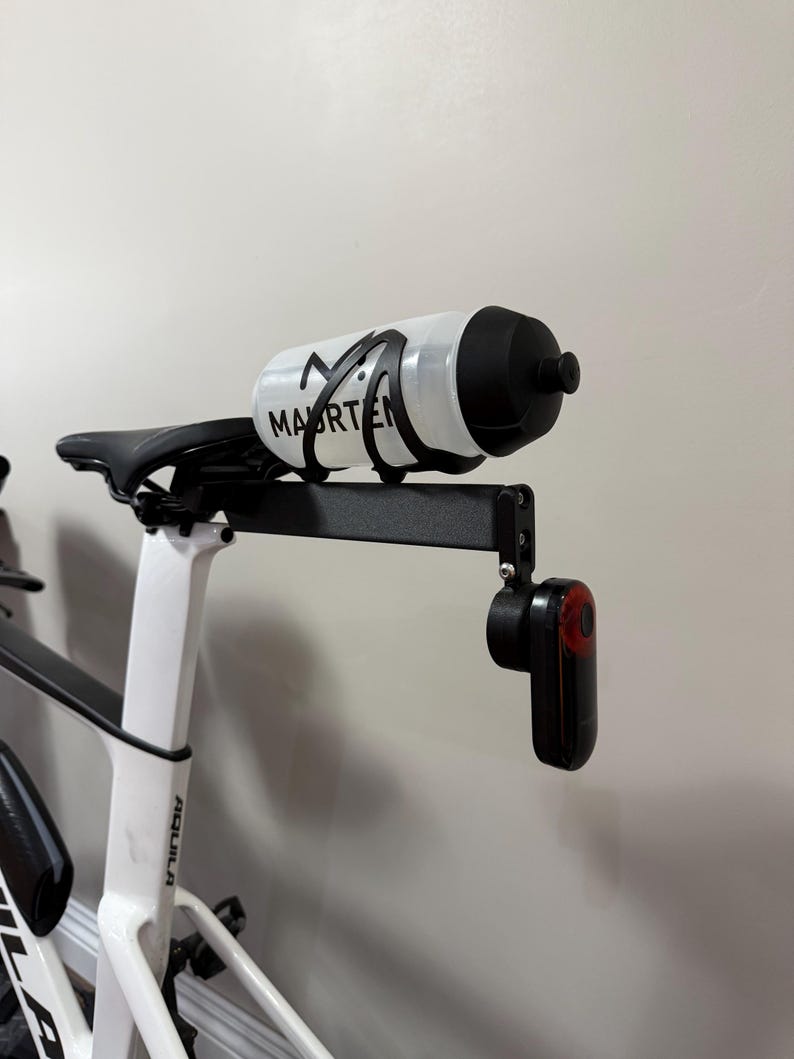 Puede incluir: Una bicicleta blanca y negra con una botella de agua y una luz trasera roja adjunta. La botella de agua tiene la palabra "MAARTEN" impresa. El asiento de la bicicleta es negro.