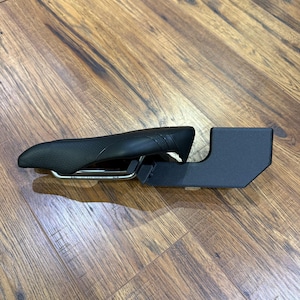 May include: A black bicycle saddle with a curved design and a silver metal frame. The saddle is connected to a black, rectangular support structure. The items are on a wooden surface.