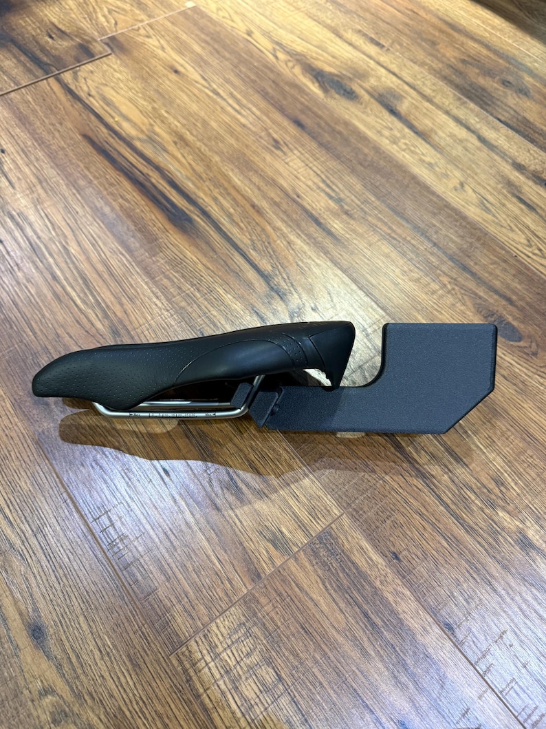 May include: A black bicycle saddle with a curved design and a silver metal frame. The saddle is connected to a black, rectangular support structure. The items are on a wooden surface.