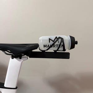 Puede incluir: Bicicleta blanca y negra con asiento negro y portabotellas. La botella de agua es blanca con texto negro "MAURTEN". La bicicleta est&aacute; montada sobre un cuadro blanco.