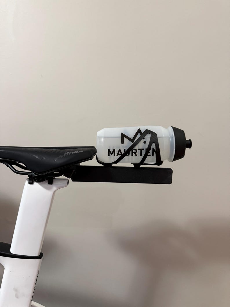 Puede incluir: Bicicleta blanca y negra con asiento negro y portabotellas. La botella de agua es blanca con texto negro "MAURTEN". La bicicleta est&aacute; montada sobre un cuadro blanco.