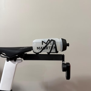 Puede incluir: Una botella de agua blanca para bicicleta con la palabra "MAURTEN" impresa en negro, montada en un soporte negro sujeto a un sill&iacute;n de bicicleta. La botella tiene una tapa negra. El sill&iacute;n de la bicicleta es negro y blanco.