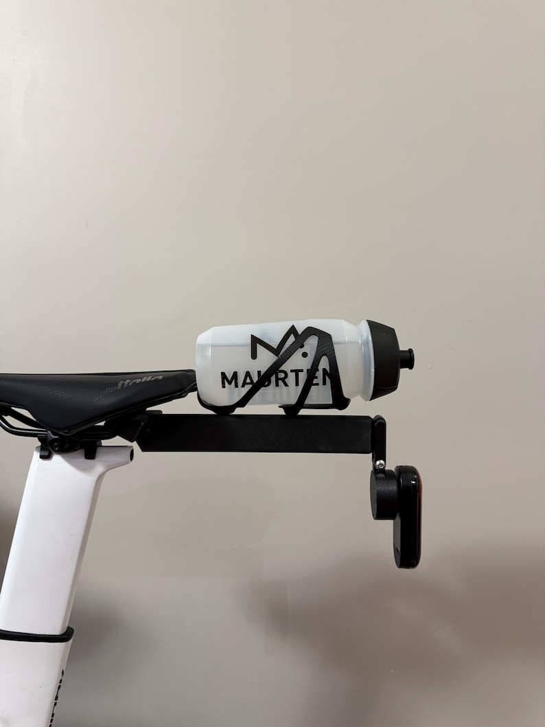 Puede incluir: Una botella de agua blanca para bicicleta con la palabra "MAURTEN" impresa en negro, montada en un soporte negro sujeto a un sill&iacute;n de bicicleta. La botella tiene una tapa negra. El sill&iacute;n de la bicicleta es negro y blanco.