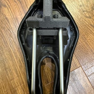 May include: A black bicycle saddle with a silver metal frame and a grey foam support. The saddle has a curved design and is mounted on a wooden surface. The saddle is designed for comfort and performance.