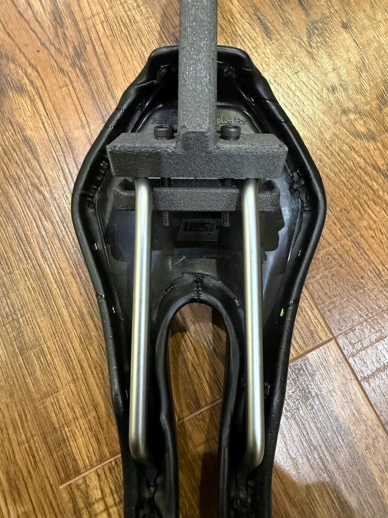May include: A black bicycle saddle with a silver metal frame and a grey foam support. The saddle has a curved design and is mounted on a wooden surface. The saddle is designed for comfort and performance.