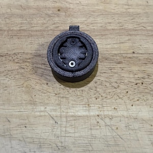 May include: A black bicycle computer mount, circular in shape. It features a textured surface and a central recess for a device. A small, silver screw is visible in the middle.