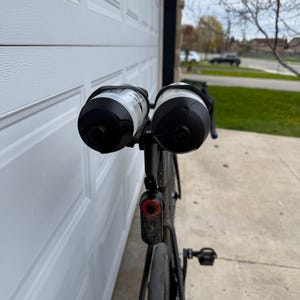 May include: Black road bike with two water bottles attached. The bottles are black and white. A red taillight is mounted below the seat. The bike is parked in front of a white garage door.