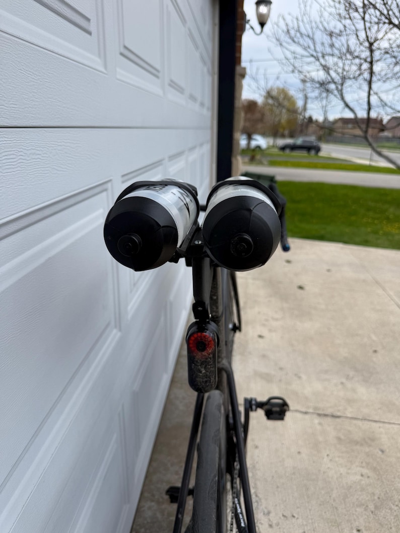 May include: Black road bike with two water bottles attached. The bottles are black and white. A red taillight is mounted below the seat. The bike is parked in front of a white garage door.