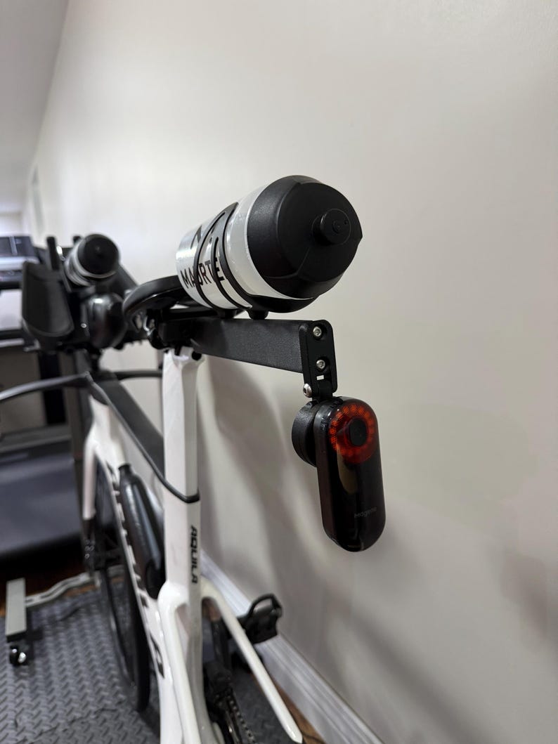 Puede incluir: Una bicicleta de carretera blanca con una extensi&oacute;n de manillar negra que sostiene una botella de agua y una luz trasera roja. La botella de agua es blanca con detalles y texto en negro. La luz trasera es negra con luz roja.