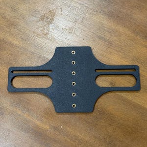 May include: A black, textured metal bracket with two rectangular cutouts and seven evenly spaced holes. The bracket is centered on a wooden surface.