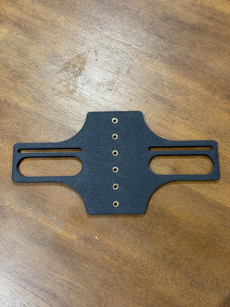 May include: A black, textured metal bracket with two rectangular cutouts and seven evenly spaced holes. The bracket is centered on a wooden surface.