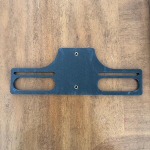 May include: A black, rectangular bracket with two oval cutouts and two small holes. The bracket has a curved top and is made of a textured material. The item is laying on a wooden surface.