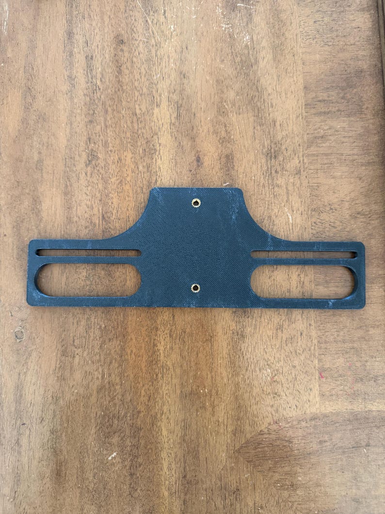 May include: A black, rectangular bracket with two oval cutouts and two small holes. The bracket has a curved top and is made of a textured material. The item is laying on a wooden surface.